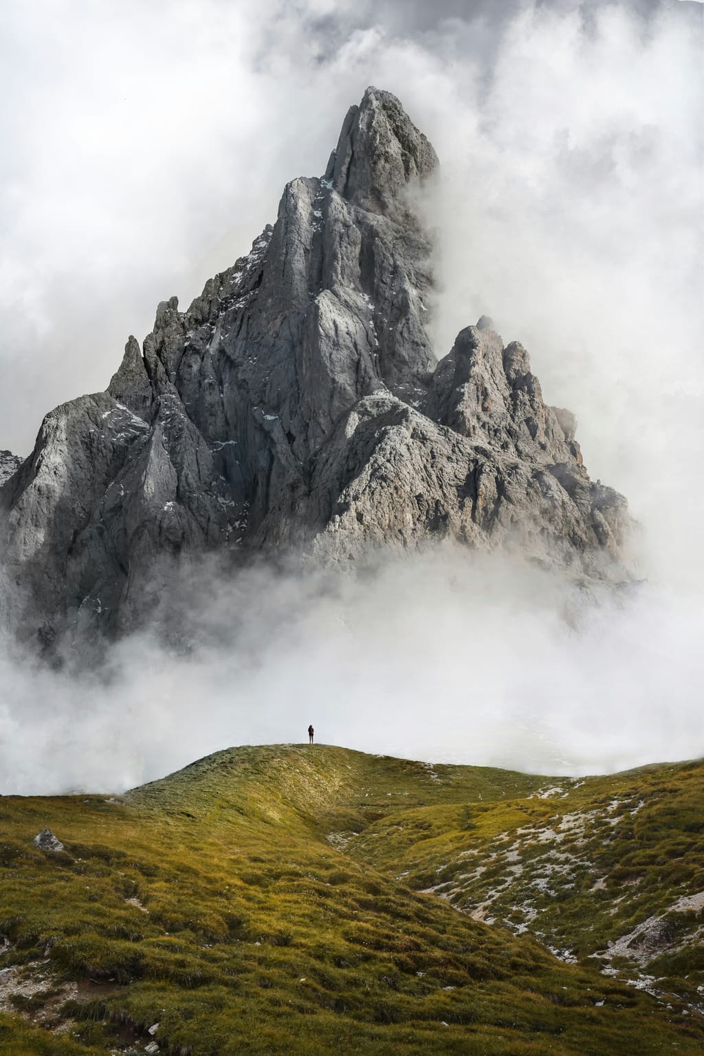 A lone hiker on a green ridge below a rocky peak in the clouds